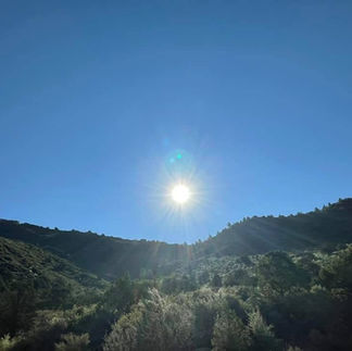 A landscape photograph of a bright, radiant sun shining directly over lush, green hillsides against a vibrant blue sky in Arizona