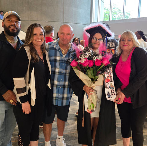 A joyful group photo celebrating a graduation. In the center, a young graduate wears a black cap and gown, a decorated graduation cap, and holds a large bouquet of pink roses. Surrounding her are smiling family members, including Michele Taft on the right wearing a vibrant pink top and black cardigan, along with Martin Taft and other loved ones indoors.