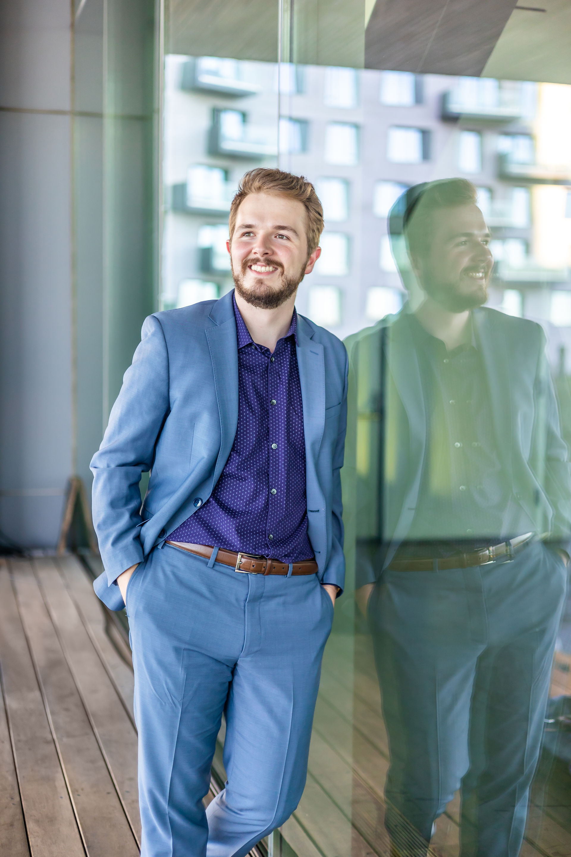 White man wearing a blue suit and purple shirt leaning against a glass wall