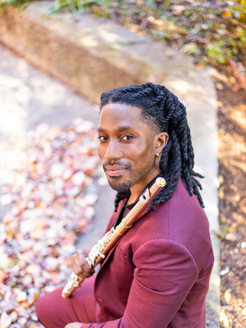 A man with dreadlocks wearing a maroon suit holding a gold flute over his left shoulder while sitting on a cement bench in Boston.