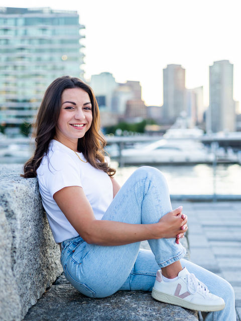 Young woman wearing a white t-shirt, light blue jeans, and sneakers, sitting on a granite bench with her leg bent in her hands in the Boston Seaport.