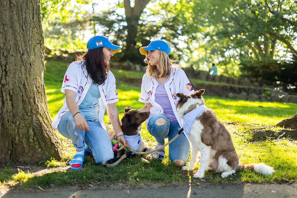 Two women wearing Red Socks jersey's, blue hats, blue jeans, and sandals posed along side their two Australian Shepherd Border Collie mix dogs