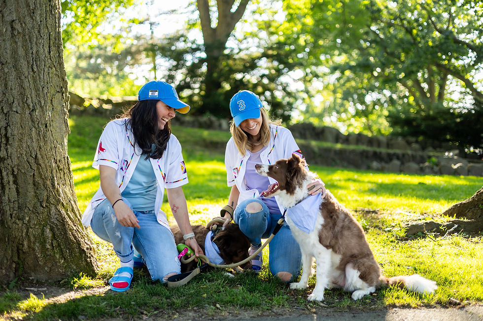 Two women wearing Red Socks jersey's, blue hats, blue jeans, and sandals posed along side their two Australian Shepherd Border Collie mix dogs