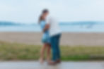 Couple kissing on a beach, the woman in a blue dress, the man in a white shirt and blue pants. Boats are in the distant water.