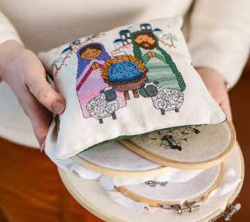 Thumbnail: Nativity cross stitch pattern in greens, purples and pinks held in with cross stitch supplies