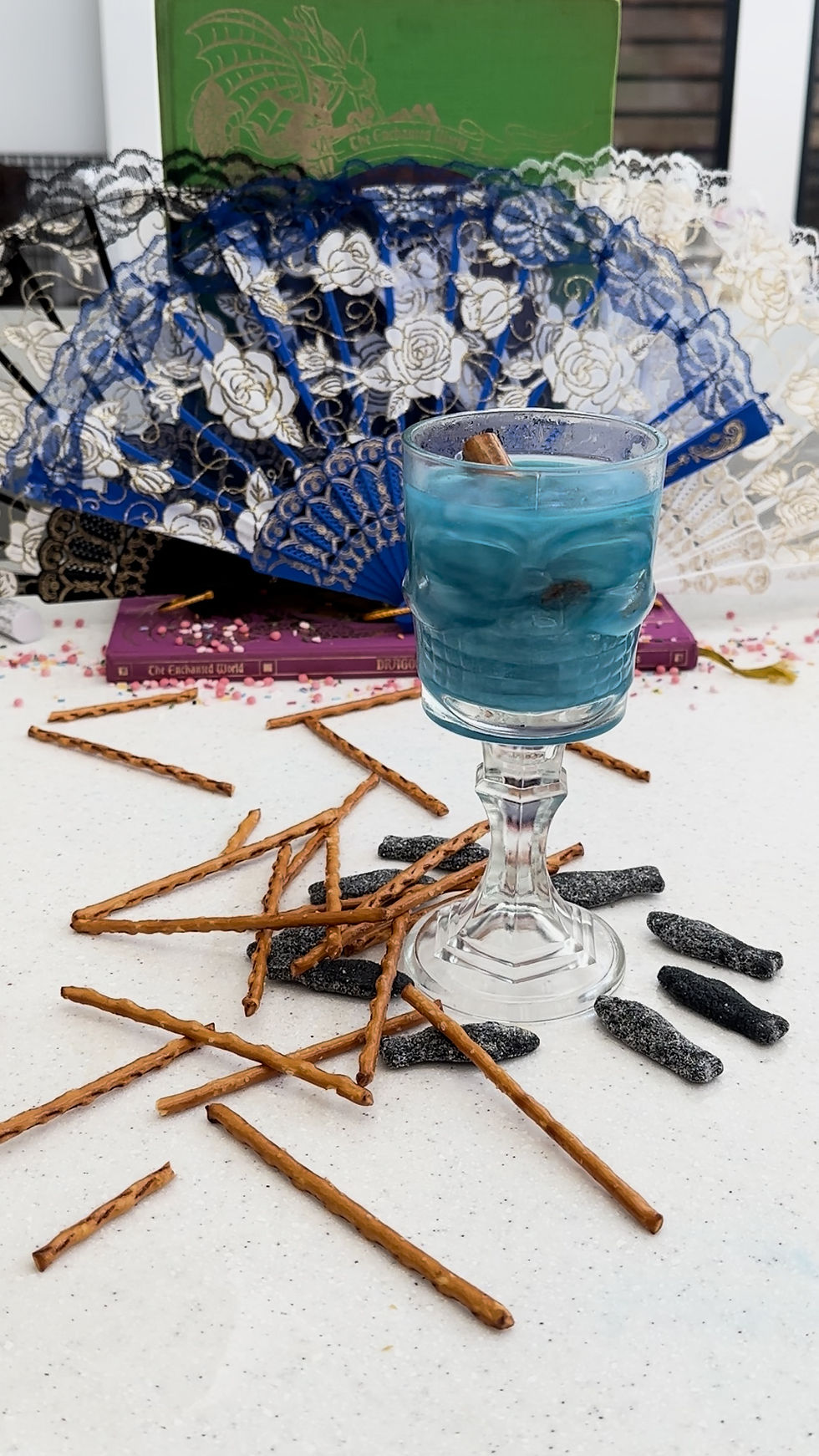 A blue drink in a glass with a cinnamon stick, surrounded by pretzel sticks and licorice, set on a table. A lacy blue fan and books in the background.