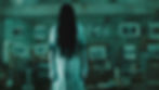 A figure in a white dress with long black hair stands ominously in a dim room with photos on the walls, creating a haunting atmosphere.
