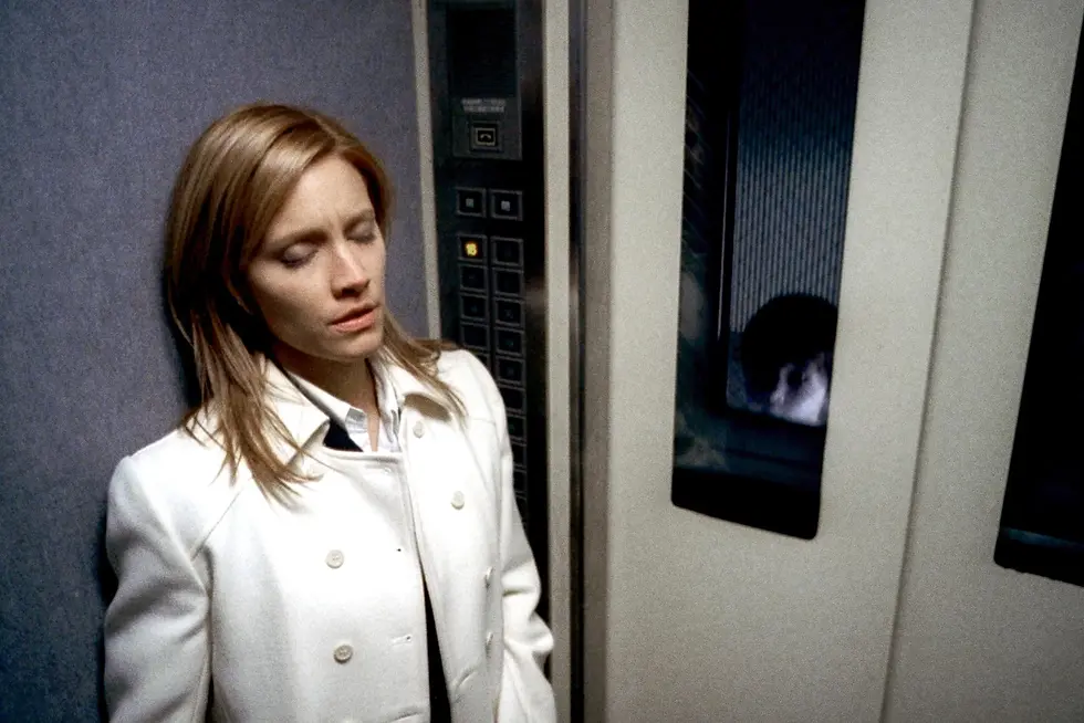 A woman in a white coat leans against an elevator wall with eyes closed, looking pensive. Elevator panel visible; reflections on the doors.