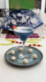 Blue cocktail with a candy garnish on a ceramic plate with small pastries. Lacy fans and books in the colorful background.
