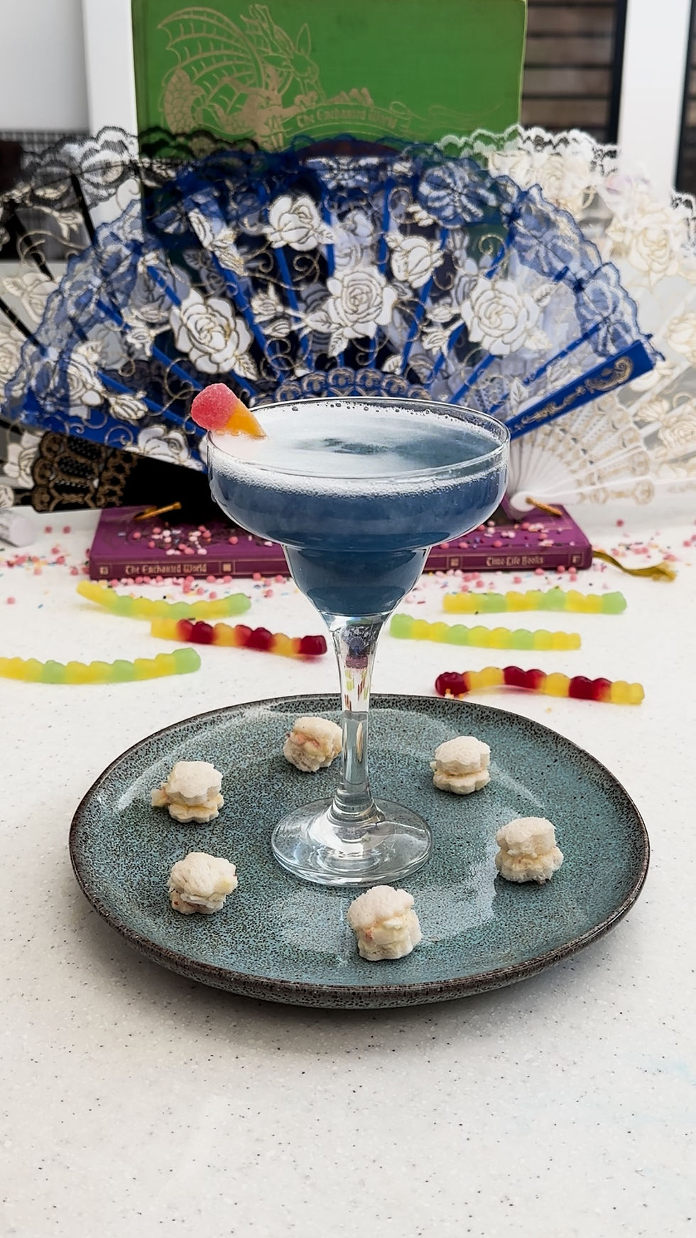 Blue cocktail with a candy garnish on a ceramic plate with small pastries. Lacy fans and books in the colorful background.