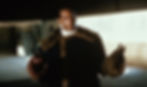 Man with a hook hand in a fur-collared coat gestures in a dimly lit space. Background shows a wall with faint text and a moody atmosphere.