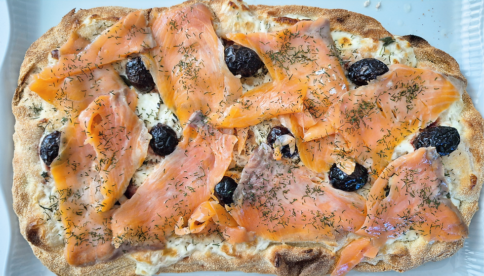 Pinsa cuite garnie de crème fraîche, d’olives noires, herbes fraîches et aneth, tranches de truite fumée, le tout posé sur une plaque de four recouverte de papier sulfurisé, vue de dessus.