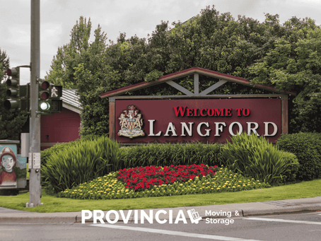 Welcome sign for Langford at a landscaped roadside corner, featuring red flowers, green shrubs, and a wooden structure, with a traffic light and crosswalk visible on an overcast day.