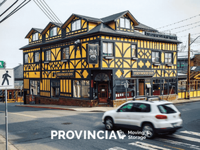 Historic yellow-and-black half-timbered Fernwood Inn building on a street corner in Fernwood, Victoria, BC, with a white SUV driving past and crosswalk and street signs in the foreground.