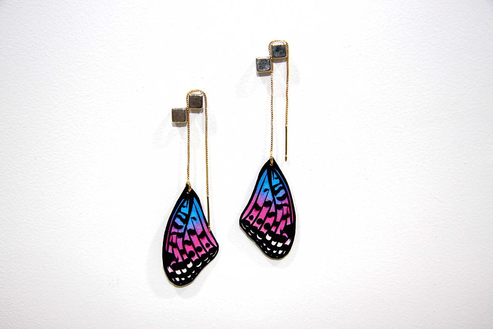 Thumbnail: Transgender Pride Flag Fritillary Butterfly Wing Earrings in 18k gold plated threads magnetized to a white Wall.