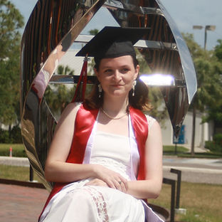 Image of a young woman in a white dress, graduation cap, and red and lavender stoles sitting on the base of a dna statue