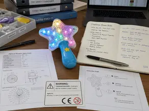 Toy product-development desk showing a light-up sample, optical module datasheet, warning draft, and compliance review notes.