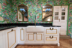 Custom white kitchen cabinets by Adam Gibbons with a farmhouse sink, black marble countertop, and two ornate gold mirrors against green floral wallpaper.