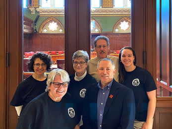 Group photo of six women and one man in front of a legislative chamber