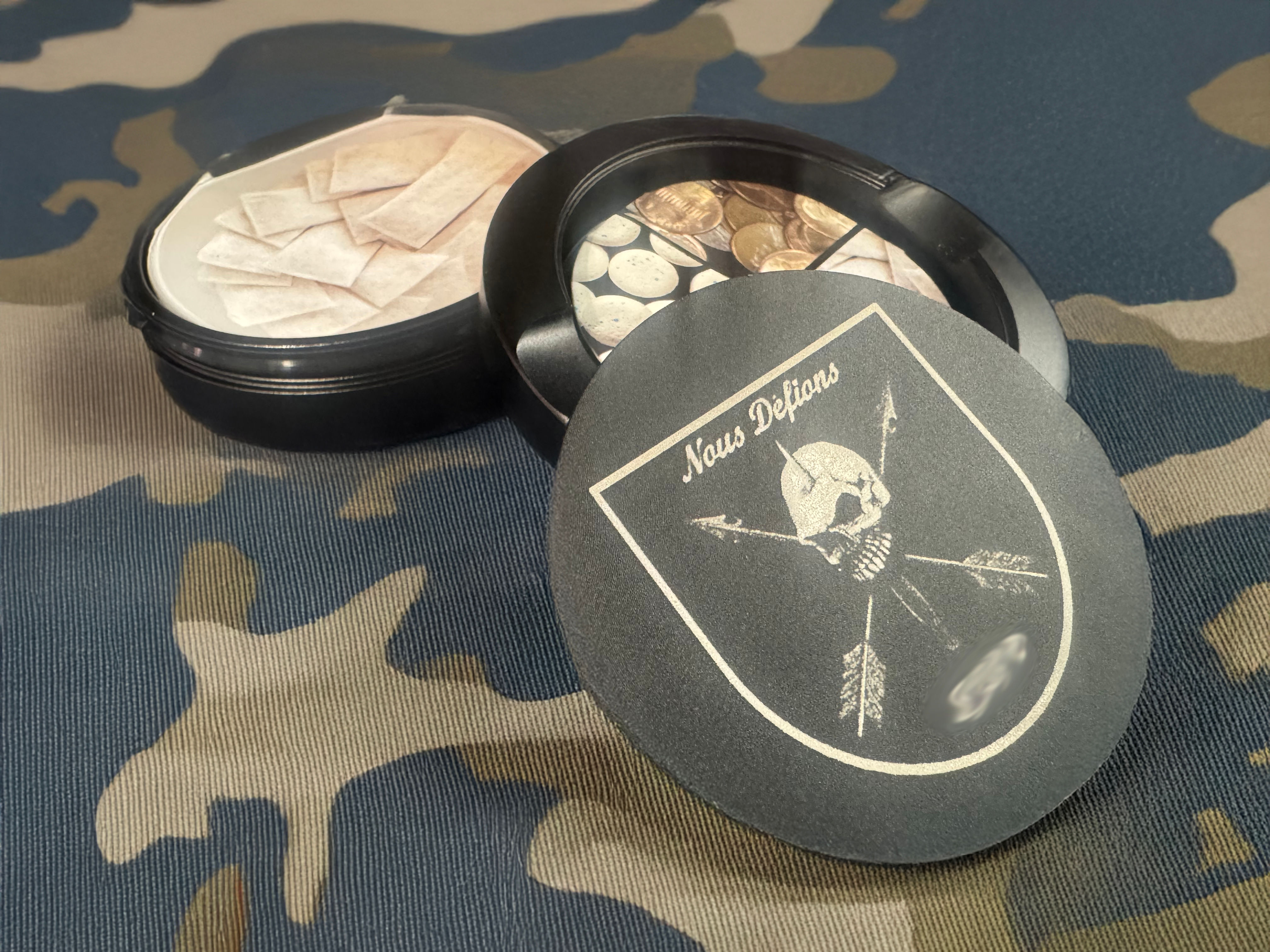 Black aluminum snus can with 'Nous Defions' skull engraving, open to pouches and coins, on camo fabric
