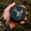 Thumbnail: Hunter holding black engraved snus can with elk head design, dew drops on lid, camo sleeve visible
