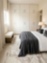 Quarto de casal decorado com estética minimalista e tons neutros. No centro, uma cama de casal arrumada com lençóis brancos, travesseiros escuros e uma manta cinza texturizada nos pés. Ao fundo, um armário embutido planejado de cor off-white preenche toda a parede, com puxadores lineares metálicos. À esquerda, uma janela com cortinas claras de tecido fluido e um banco estofado em bouclé branco com base de madeira. No piso, destaca-se um grande tapete felpudo com padronagem abstrata em tons de bege e marrom claro, e à direita, um abajur com base dourada aceso sobre a mesa de cabeceira.