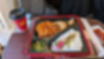 A bento box with fried cutlet, rice, and vegetables. A red "HINOTORI" cup and utensils are on a train table. Cozy travel setting.