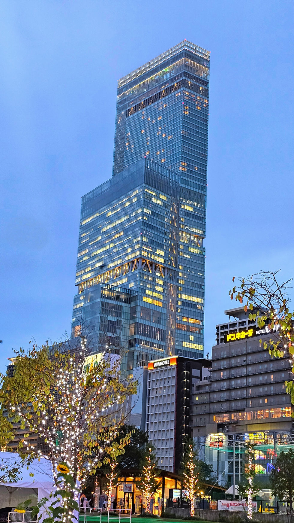 Tall glass skyscraper at dusk with lights on, surrounded by trees decorated with white lights. Signs visible on nearby buildings. Calm ambiance.