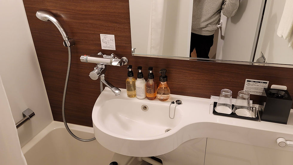 Bathroom with a white sink, four soap dispensers, mirror, and wooden wall. A person in gray sweater is partially visible in the mirror.