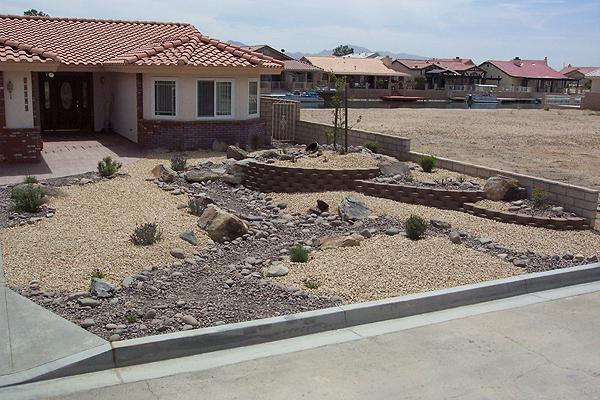  Desert home with a terracotta roof and xeriscape yard featuring rocks and sparse shrubs. A flat landscape and neighboring homes are visible.