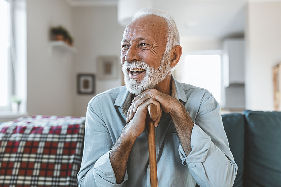 Elderly man with dementia with a white beard smiling warmly, leaning on a cane in a cozy living room with a plaid blanket and soft lighting.