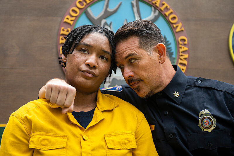 Two people stand close; one in a yellow uniform looks serious, the other in a black uniform whispers. Background shows a camp emblem.