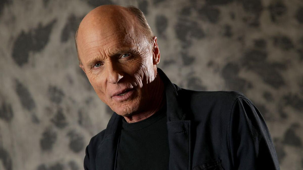Bald man in a black jacket and shirt, looking confidently at the camera. Background is abstract with dark gray patterns.