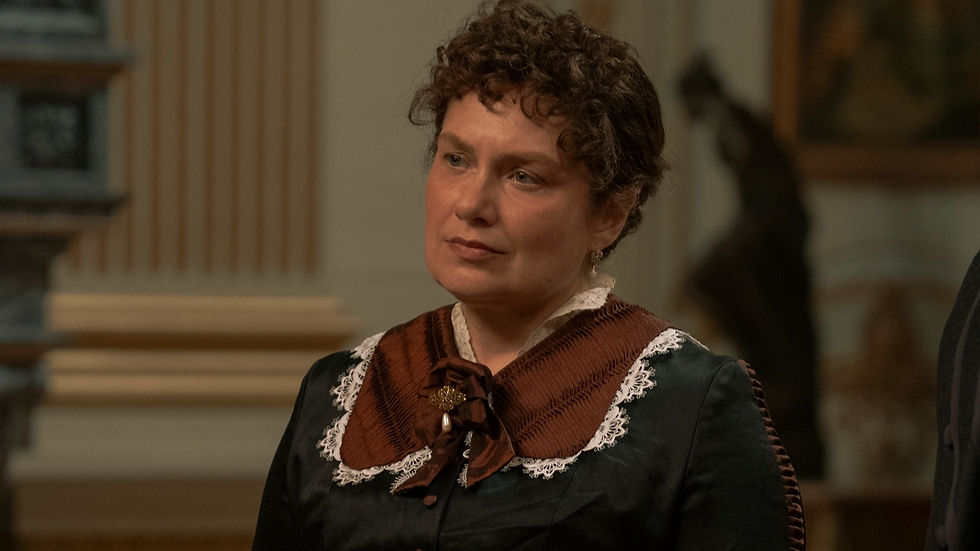 A woman in a vintage dress with lace trim and a brooch stands indoors, looking thoughtful. Ornate decor is blurred in the background.