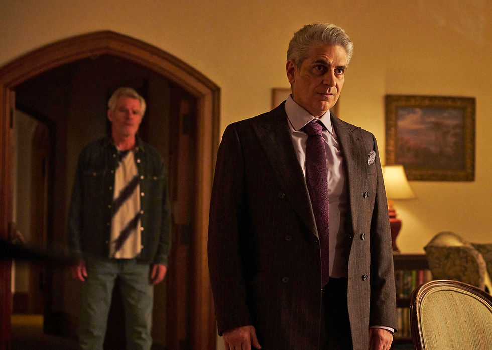 Man in suit stands with a serious expression, while another man watches from a doorway. Dimly lit room with lamp and painting in background.