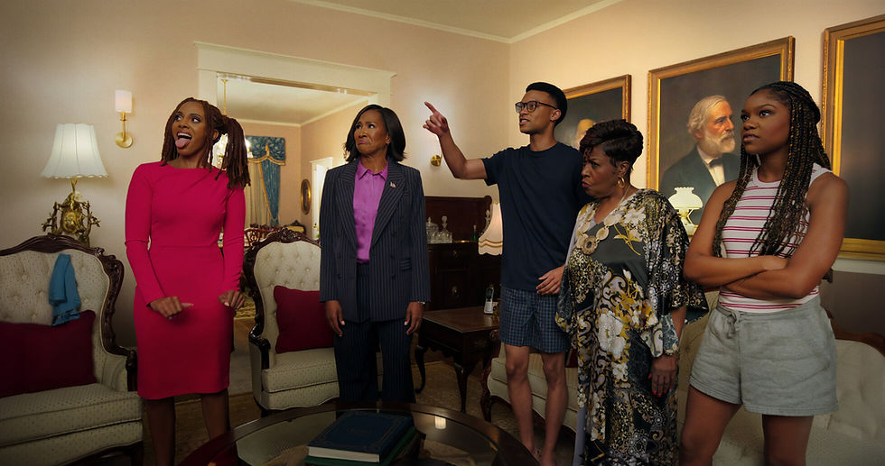 Five people in a living room; one gestures, another in pink reacts playfully. They stand near vintage furniture and large portraits on the wall.