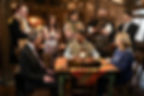 A group of eight people in varied costumes talk around a table with a lantern. They appear serious in a warmly lit, rustic room.