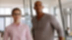Two coworkers, a woman in pink shirt and glasses and a man in gray shirt, stand in a bright office, looking serious.