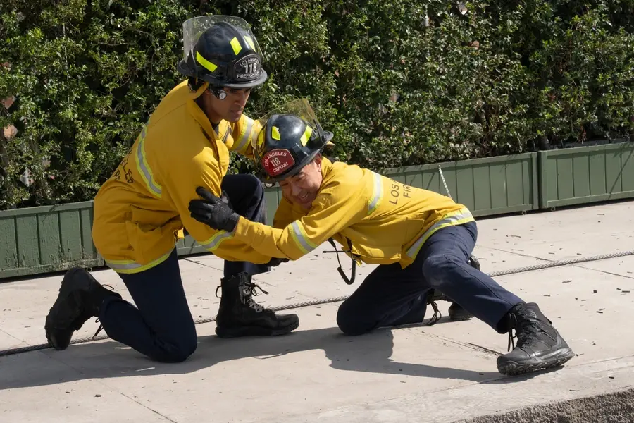 Two Los Angeles firefighters in yellow gear kneel and brace each other during a training drill on a sunlit sidewalk.