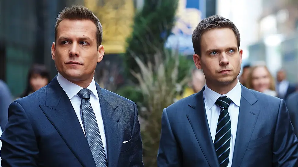 Two men in suits walk in a city setting, appearing focused. The background is blurred with people and greenery. Blue suits and striped ties stand out.
