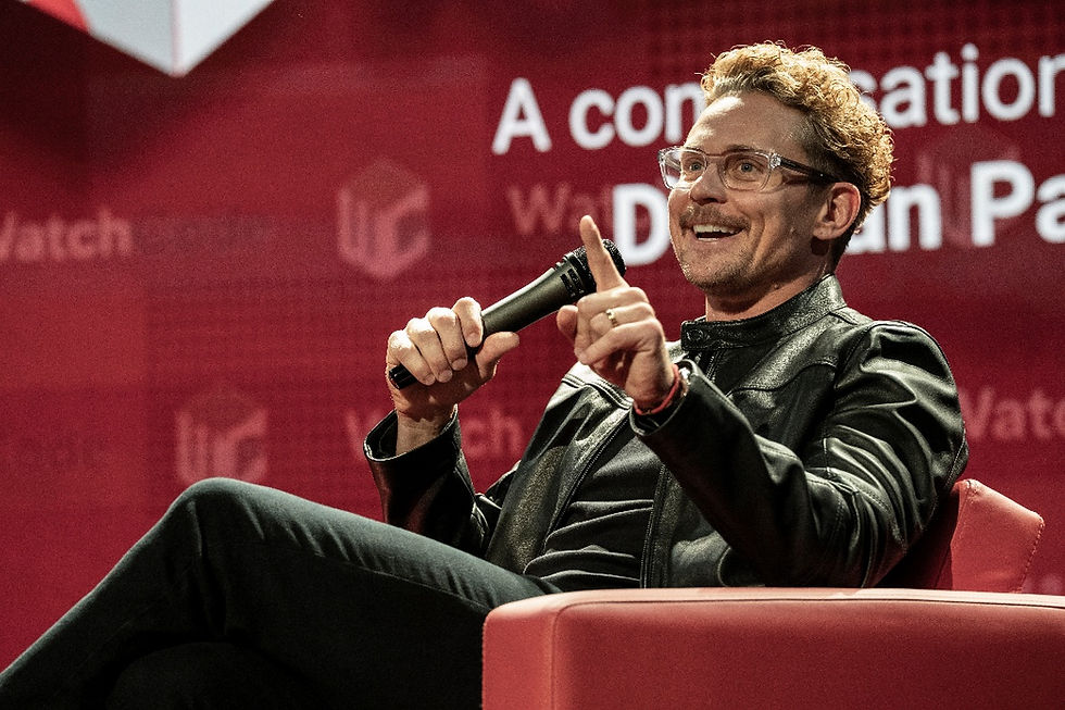 Man in glasses and black jacket speaks into a microphone, pointing on a red stage with blurred white conference text.