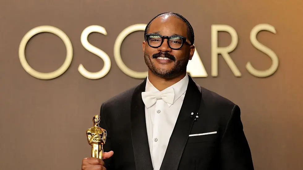 Smiling man in black tuxedo and glasses holds an Oscar statue in front of an OSCARS backdrop.