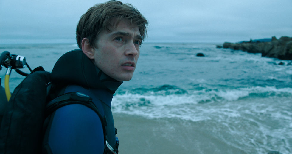 Young man in a wetsuit and scuba gear stands by a rocky ocean shore, gazing worriedly at the waves.