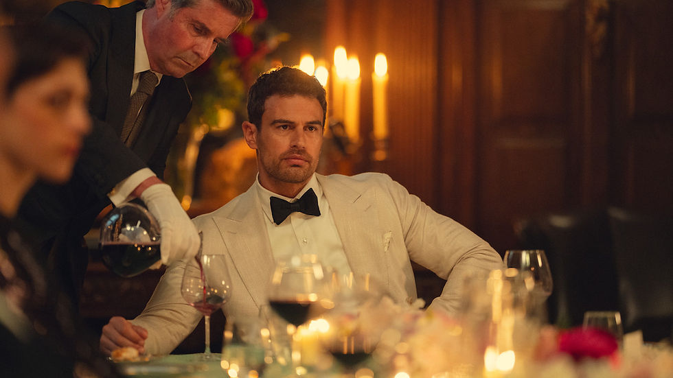 Man in white tuxedo sits at a candlelit dinner as a waiter pours wine, with blurred guests and glasses in warm light.