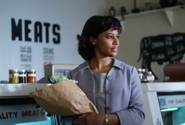 Woman holding flowers in a butcher shop, wearing a light purple coat. Meat counter and jars in the background. Calm, thoughtful mood.