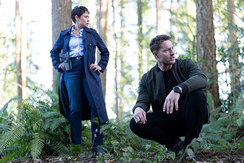 Two people in a forest, one standing with a gun, the other crouching. Both are alert, surrounded by tall trees and green ferns.