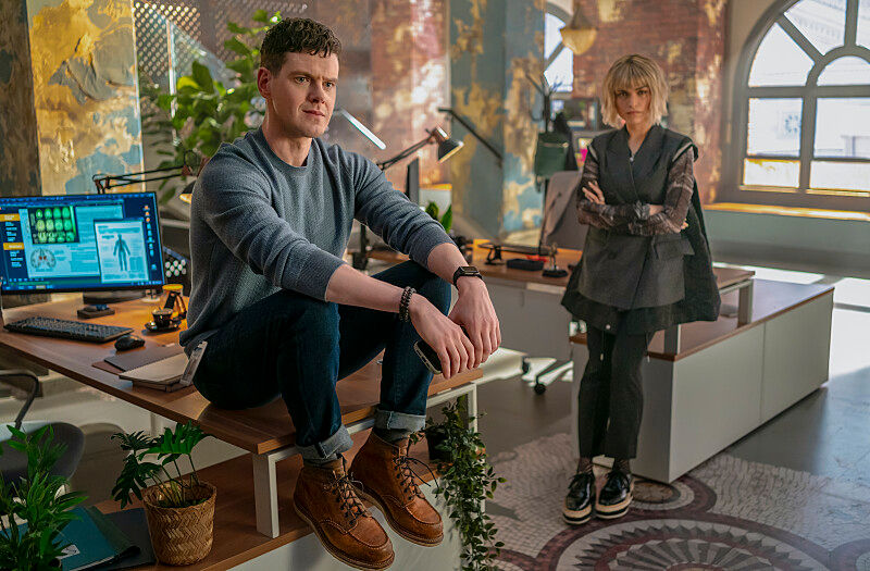 Man sitting on a desk next to a woman standing with arms crossed in a colorful office. Computers and plants are present, sunlight through arched window.