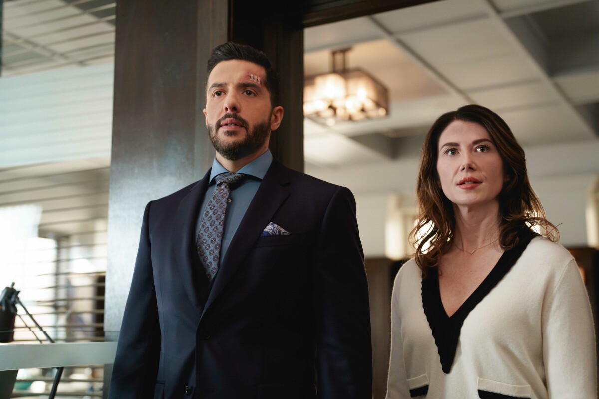 Man in a suit and woman in a white sweater stand indoors, looking surprised. Office setting with modern decor and soft lighting.