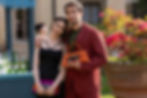 A couple poses happily; she in a black dress, he in a red suit holding an orange bag. Background: bottles, pink flowers, daylight.