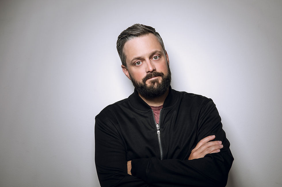 Man with a beard and short hair, arms crossed, standing against a plain white wall, wearing a black jacket and maroon shirt, neutral expression.